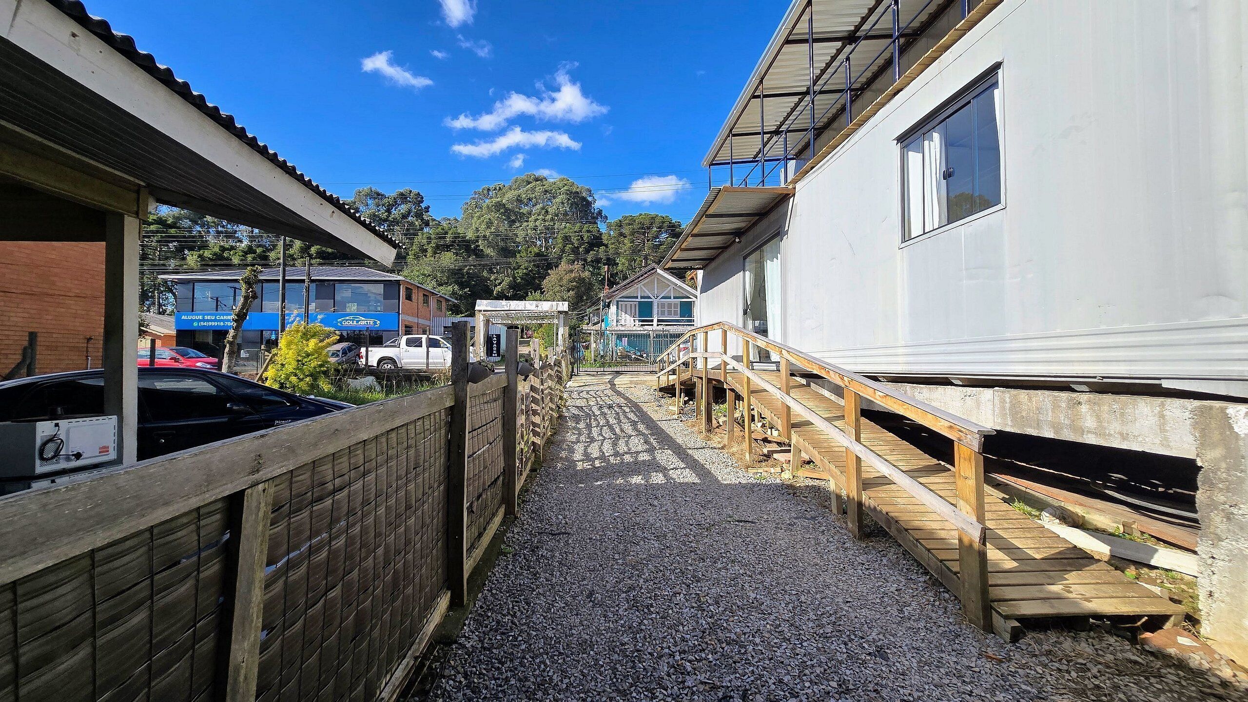 A narrow alleyway with a gravel path, wooden fence, and a wooden bridge leading to a white building with a balcony.