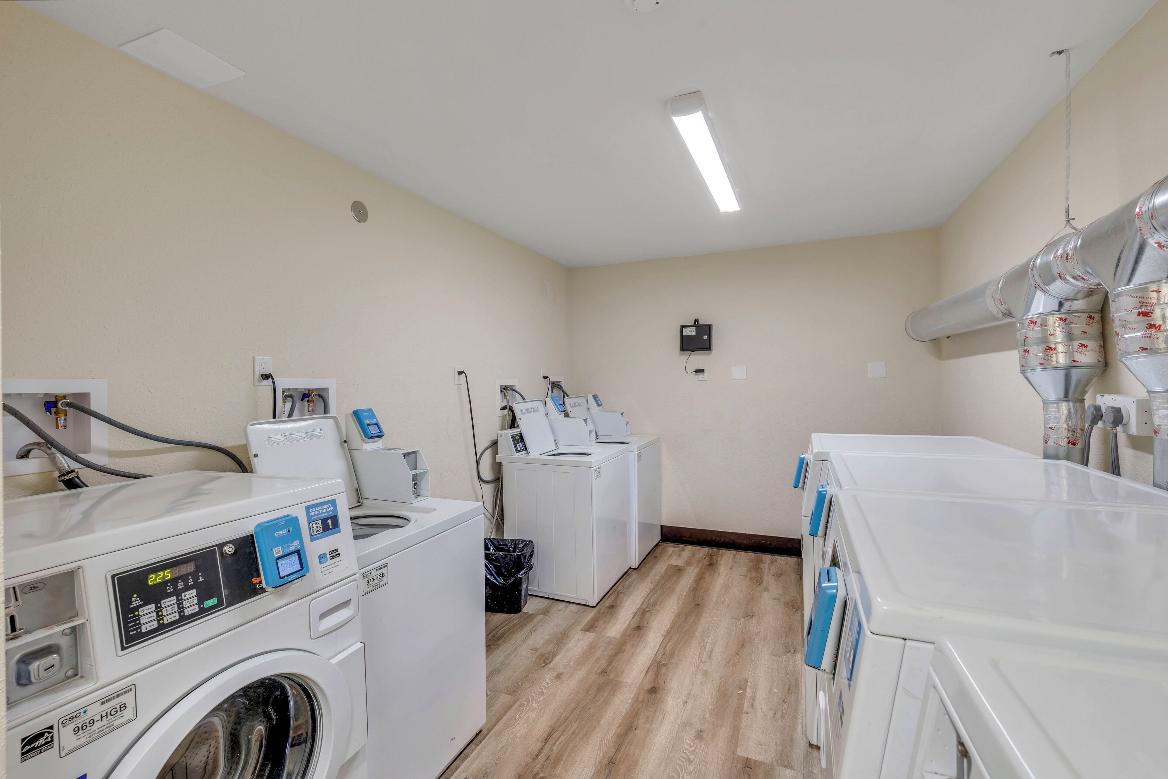 A laundry room with multiple washing machines and dryers, a trash bin, and a control panel.