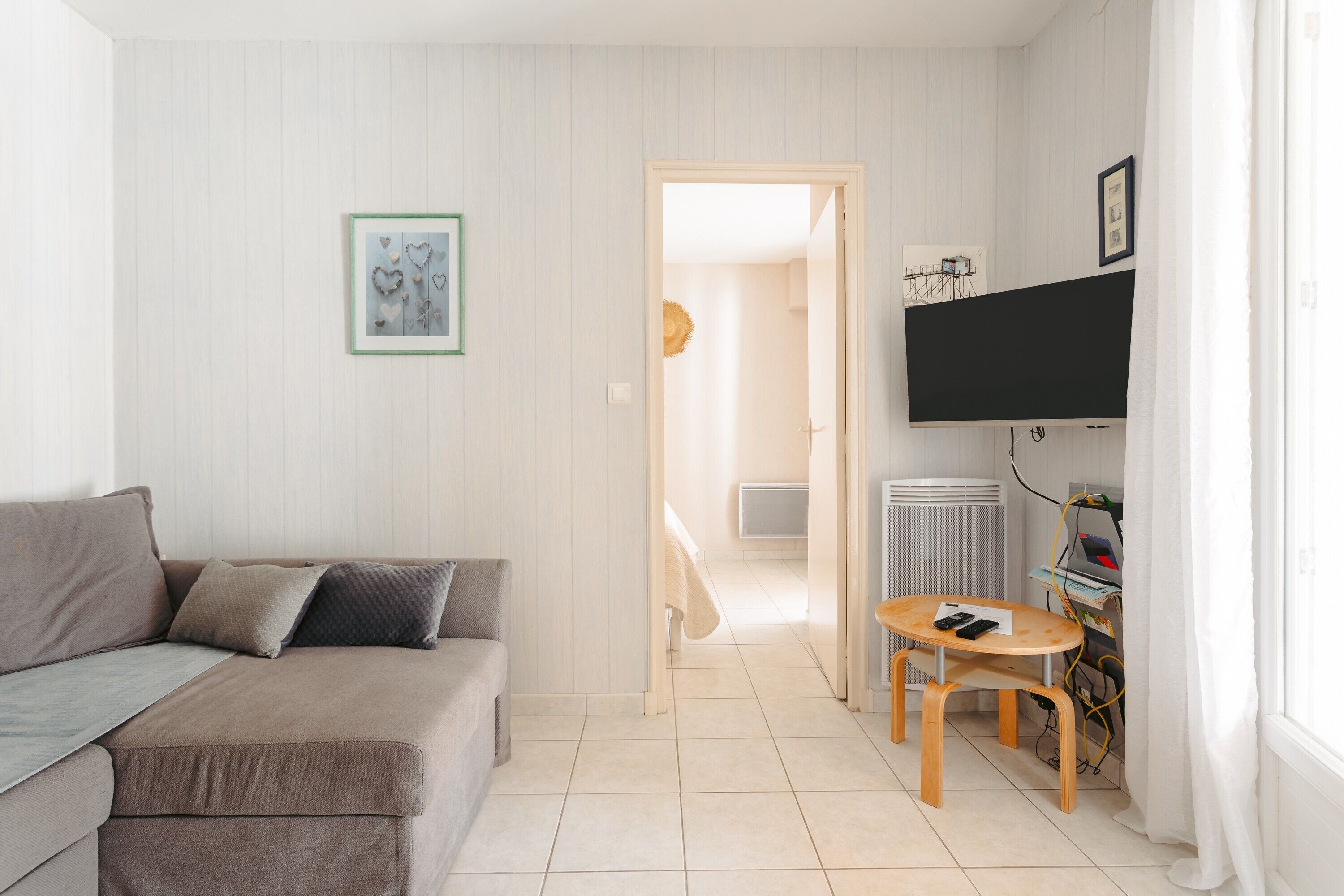 A living room with a sofa, a small round table, a TV mounted on the wall, and a door leading to another room.