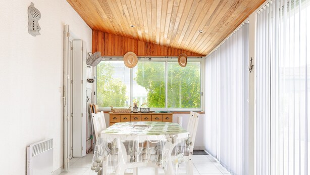 A dining area with a table covered in a patterned tablecloth, white chairs, a wooden ceiling, and large windows with blinds.