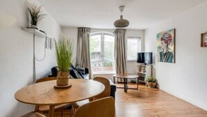 A modern living room with a round wooden dining table, a sofa, a TV, and a large window with curtains. - Central Oxford Oasis - Close to Colleges Shops (Oxford)