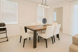 A modern dining room with a wooden table and white chairs, a decorative light fixture, and a view of the adjacent living area. - Modern 6BR Villa Heated Pool Solara Resort (Kissimmee)