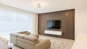 Un salon moderne avec un canapé d’angle, un téléviseur à écran plat fixé sur un mur en panneaux de bois et une grande fenêtre agrémentée de rideaux légers.