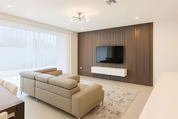 A modern living room with a sectional sofa, a flat-screen TV mounted on a wooden panel wall, and a large window with sheer curtains. - Modern 6BR Villa Heated Pool Solara Resort (Kissimmee)