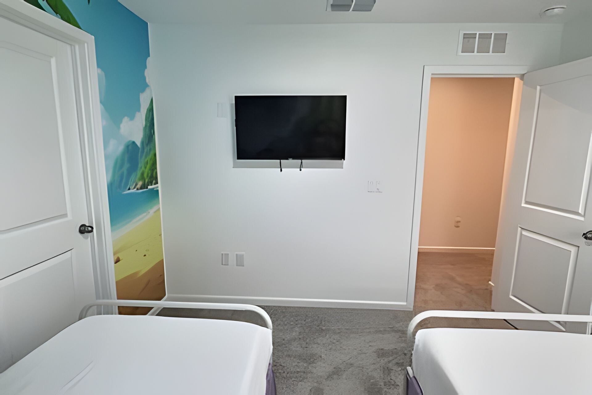 A room with two beds, a TV on the wall, and a mural of a beach scene.