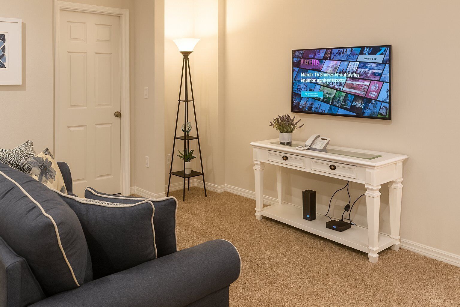 Sala de estar com sofá, mesa de console branca com telefone e uma pequena planta, luminária de chão e televisão na parede exibindo uma colagem de imagens.