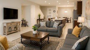 A modern living room with a grey sofa, a coffee table, and a TV. There is a kitchen in the background with a dining area. - 3bd/3.5ba Retreat With Private Pool Solara Resort (Kissimmee)