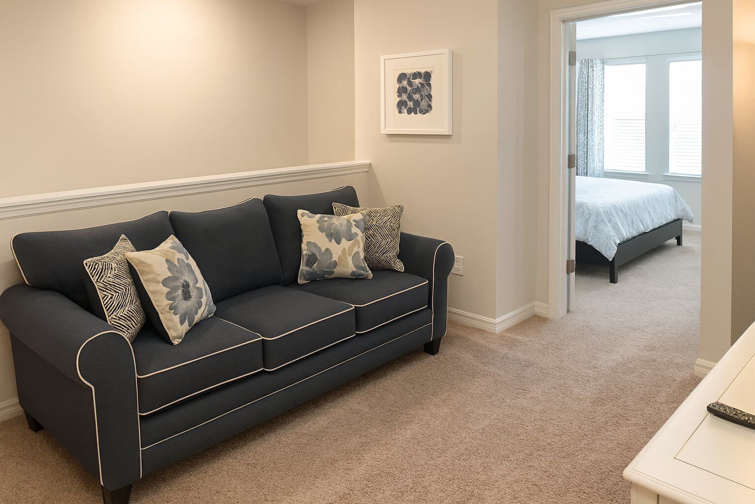 Un salon moderne avec un canapé gris foncé, des coussins à motifs, une œuvre d’art encadrée et une vue sur une chambre à coucher, visible à travers une porte ouverte.