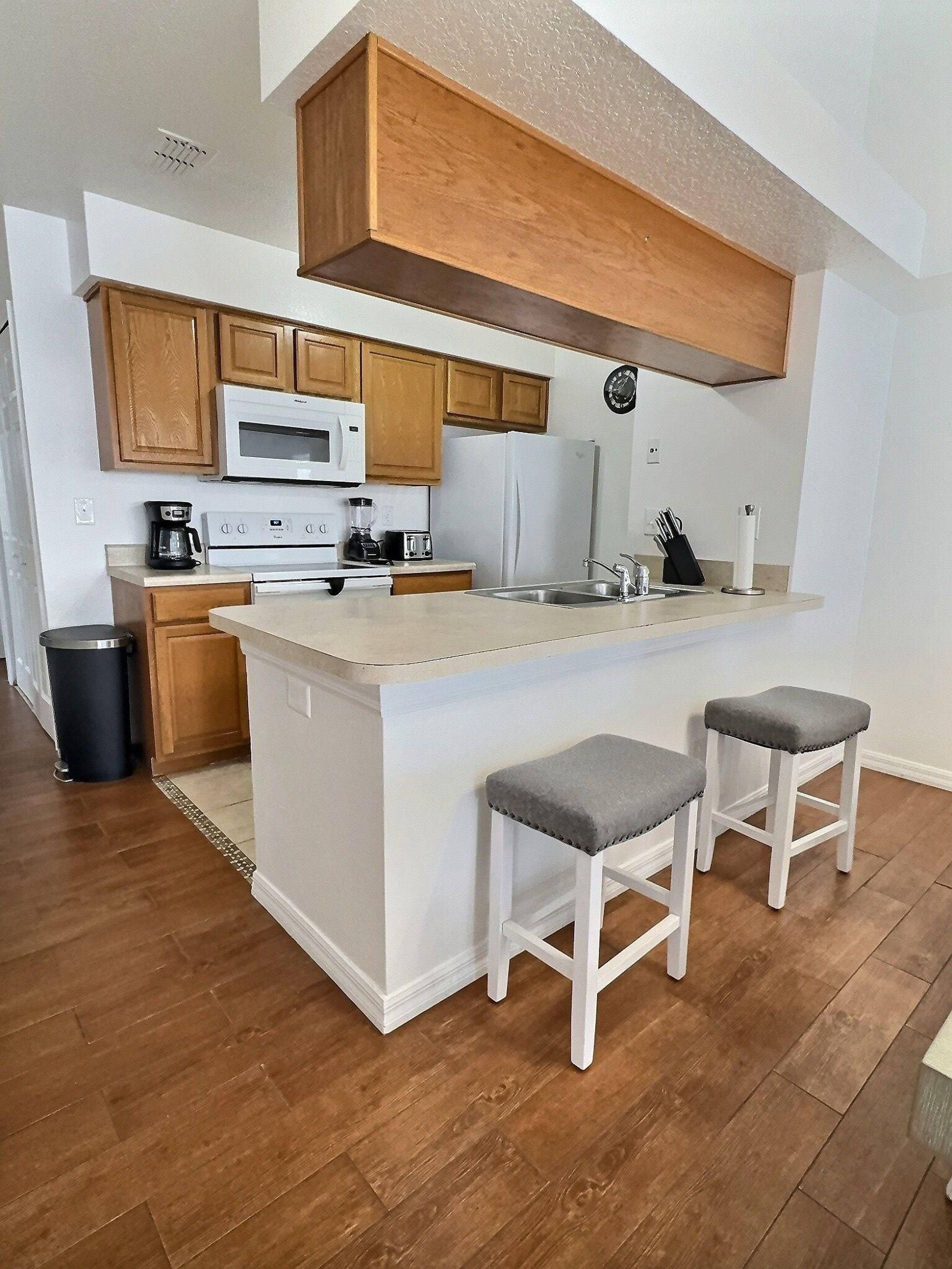 Una cocina con armarios de madera, una encimera blanca, microondas integrado, refrigerador y dos taburetes de bar.