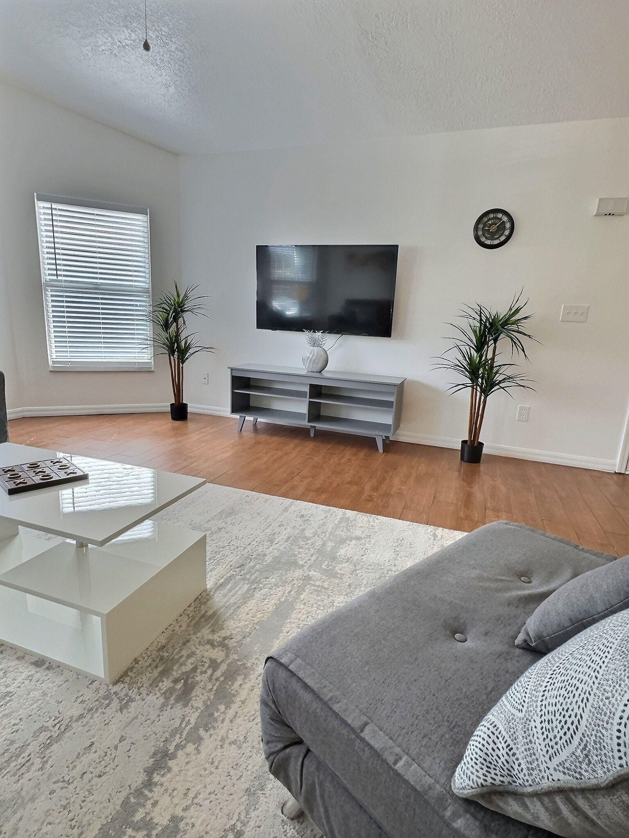 Un salon moderne avec un téléviseur à écran plat, une table basse, un canapé gris et deux plantes en pot.