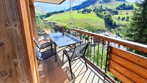 Un balcon avec une table et des chaises, une balustrade en bois et une vue sur des collines verdoyantes et des montagnes.