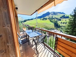 A balcony with a table and chairs, a wooden railing, and a view of green hills and mountains.