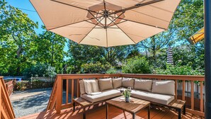 A wooden deck with a beige sectional sofa, a small table, and a large umbrella providing shade.