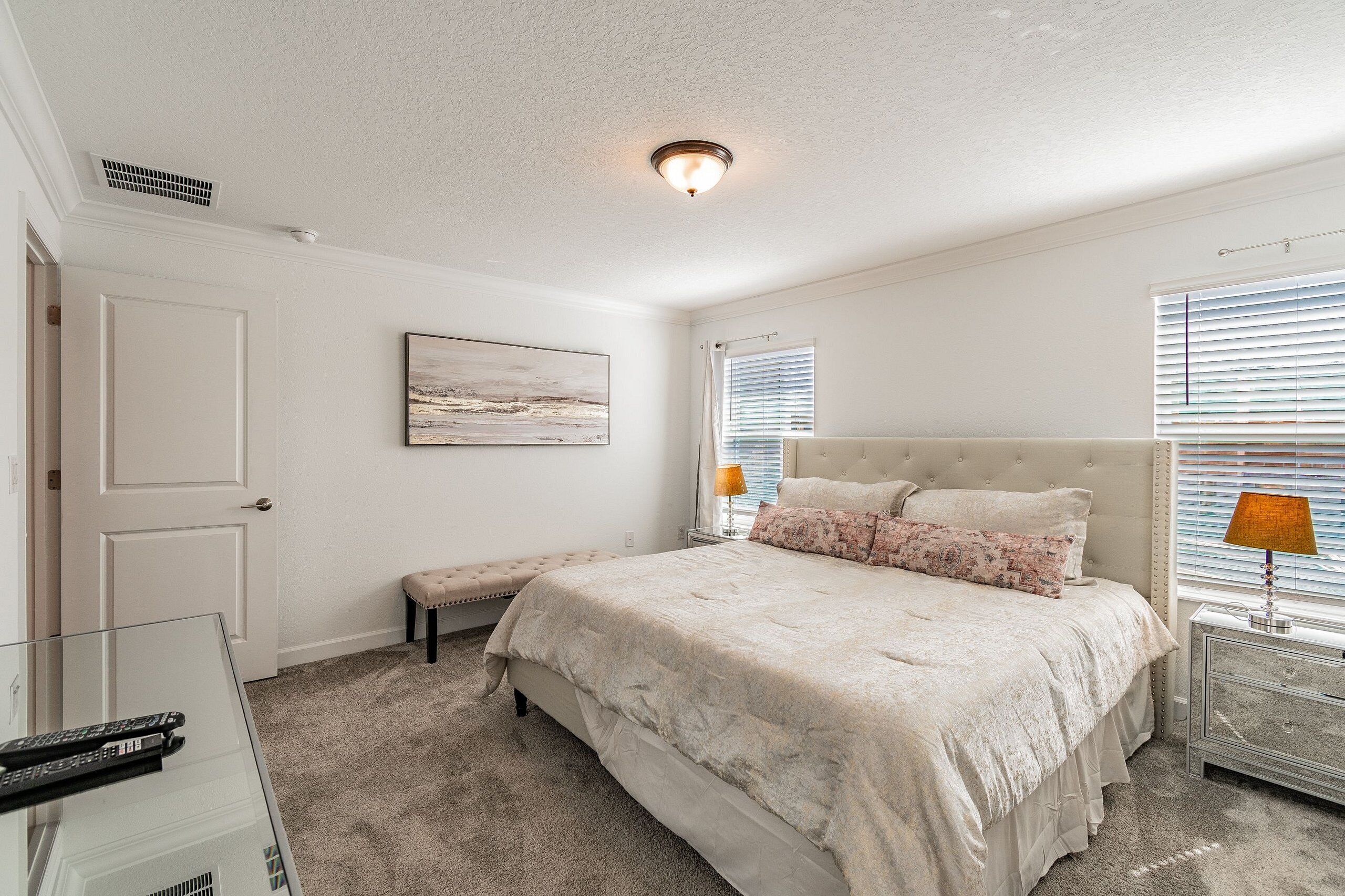 A bedroom with a large bed, a bench, a bedside table with a lamp, a painting on the wall, and a door.