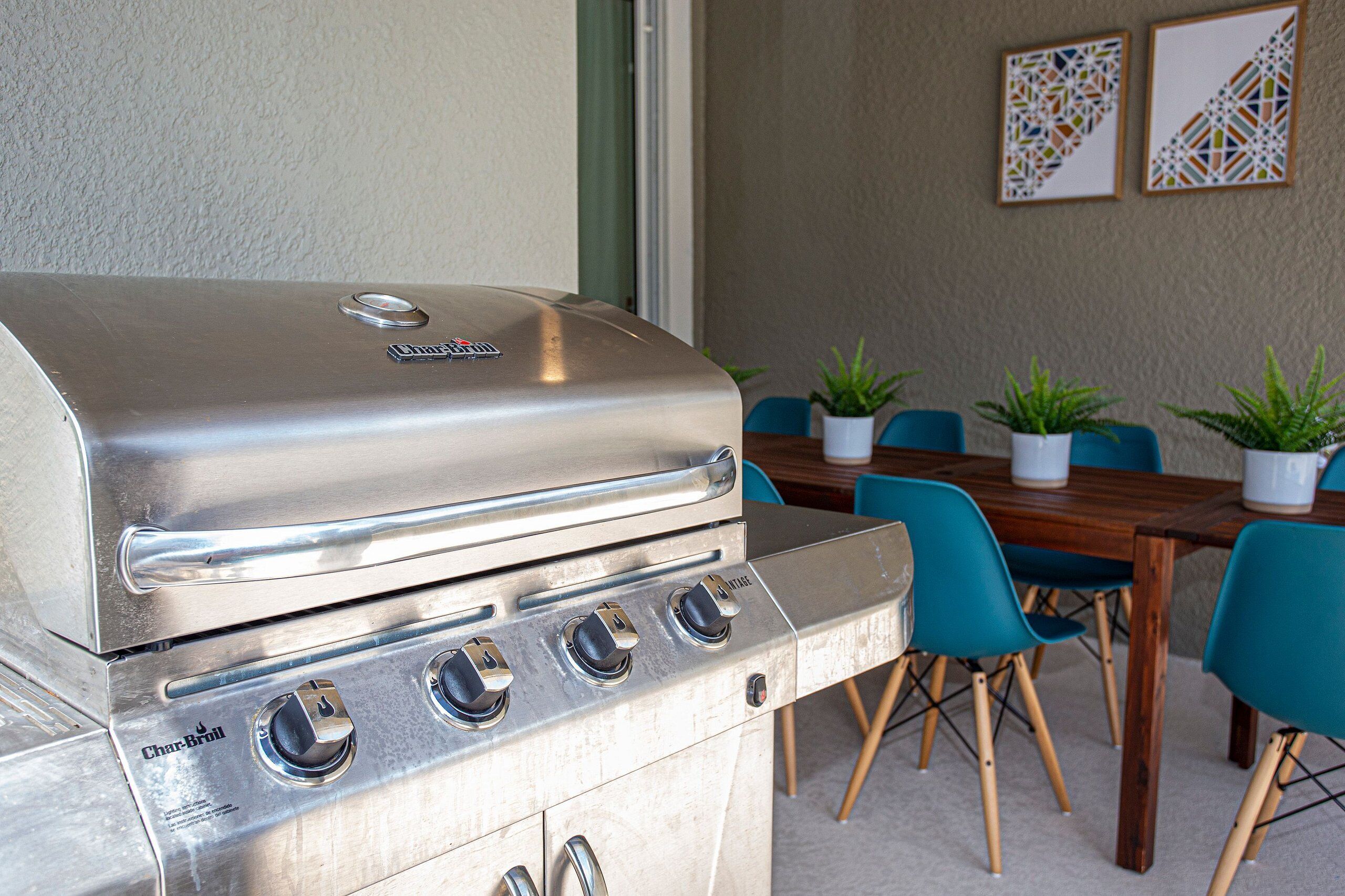 Una parrilla de gas Char-Broil de acero inoxidable con cuatro quemadores, un panel de control y una tapa. Al fondo, se ve una mesa de comedor de madera con sillas azules y plantas en macetas.