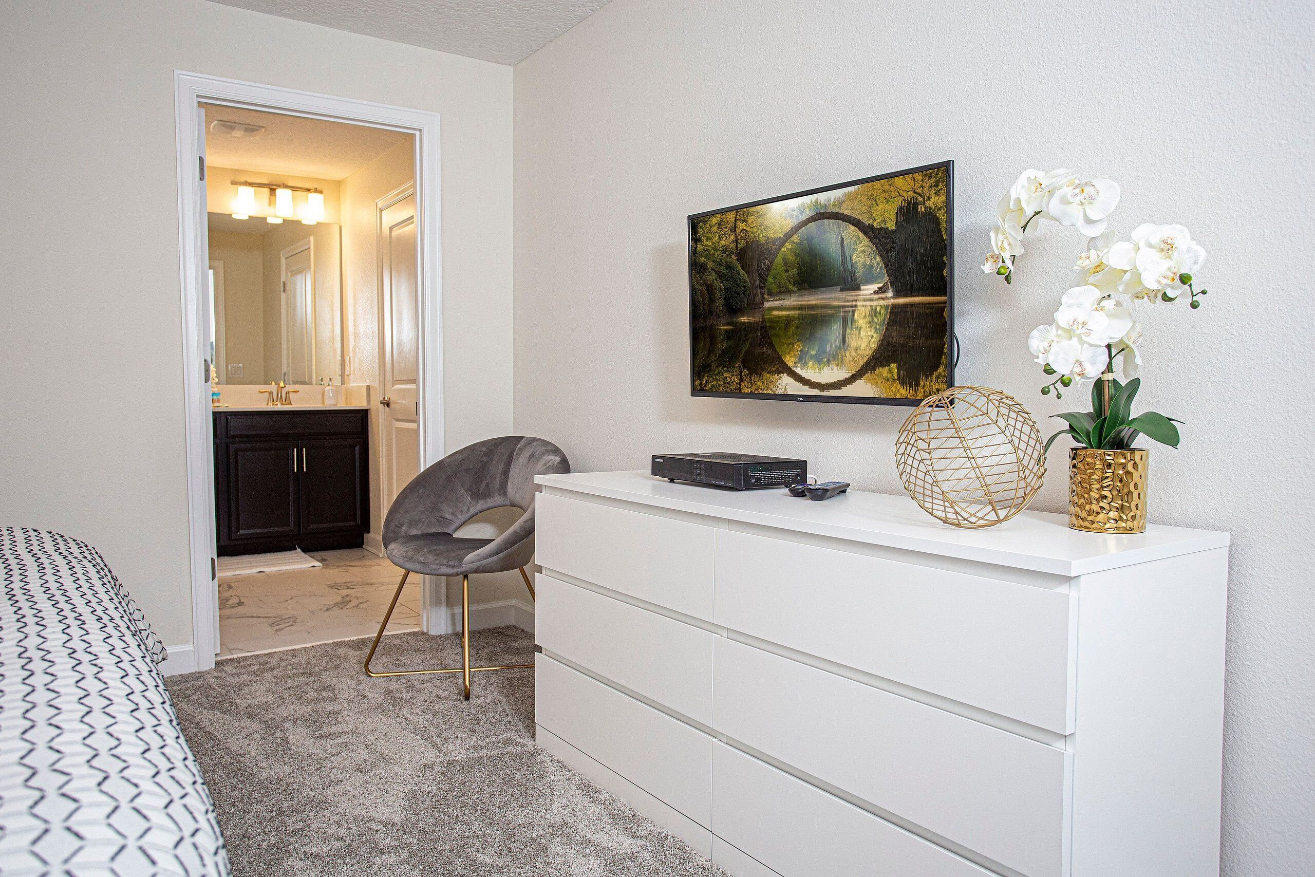 A bedroom with a bed, a dresser, a TV mounted on the wall, and a door leading to a bathroom.