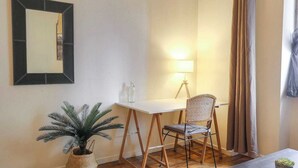 A small room with a white desk, a wicker chair, a potted plant, a lamp, and a mirror on the wall. - Charming with balcony in the heart of the 7th (Lyon)