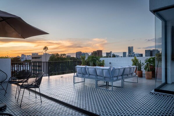 A rooftop terrace with a city view at sunset, featuring outdoor seating and potted plants. - Loft con balcón en la Americana | Lapso at Alarcón (Guadalajara)
