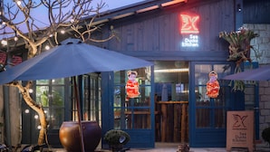 Un restaurant avec une enseigne au néon, des tables en terrasse et un grand bac à fleurs.