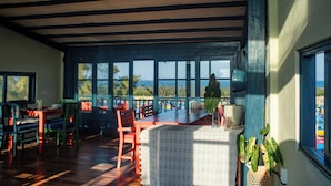 Un espace repas avec des tables et des chaises en bois, de grandes fenêtres donnant sur la mer et une plante en pot posée sur la table.