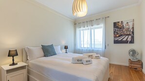 Een hotelkamer met een groot bed, twee nachtkastjes met lampen, een raam met dunne gordijnen, een schilderij aan de wand en een staande ventilator.