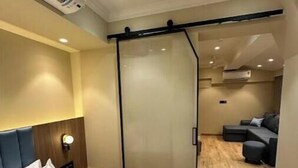 Una habitación de hotel con cama, mesita de noche, lámpara fijada a la pared y puerta corrediza de vidrio que da a una sala de estar con sofá y mesa de centro.