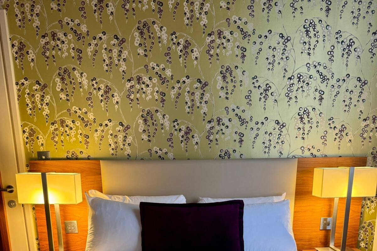 A neatly made bed with white linens and a dark purple pillow, flanked by two bedside tables with lamps, in a room with floral wallpaper.