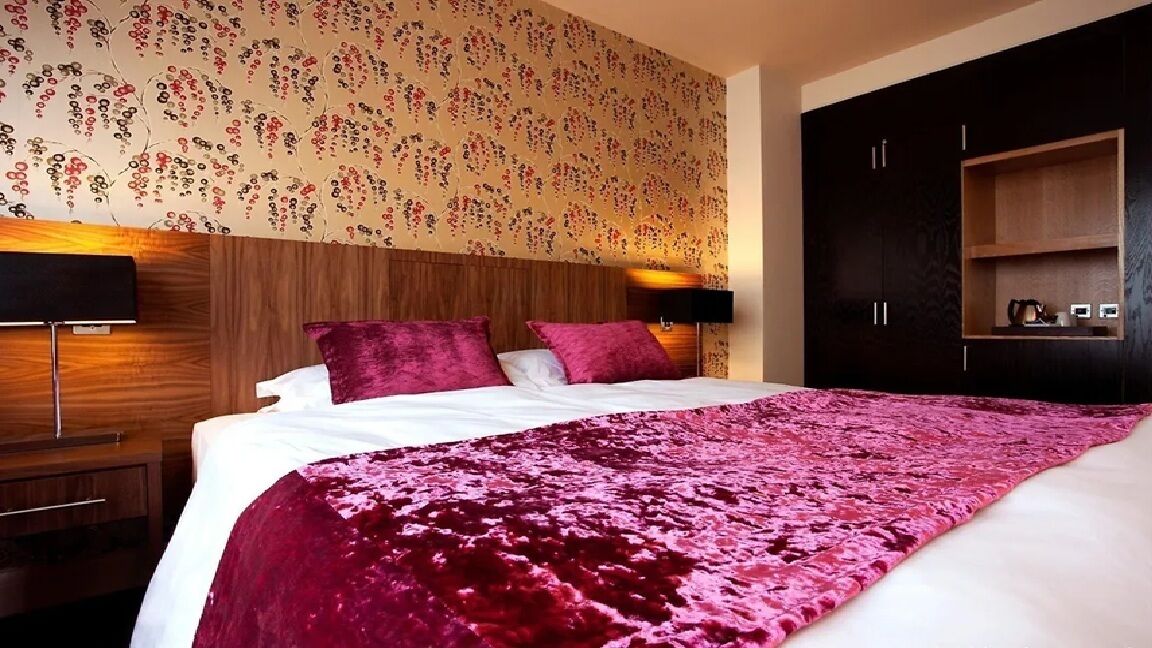 A hotel room with a large bed, two pillows, a wooden headboard, a nightstand with a lamp, a wardrobe, and a floral wallpaper.