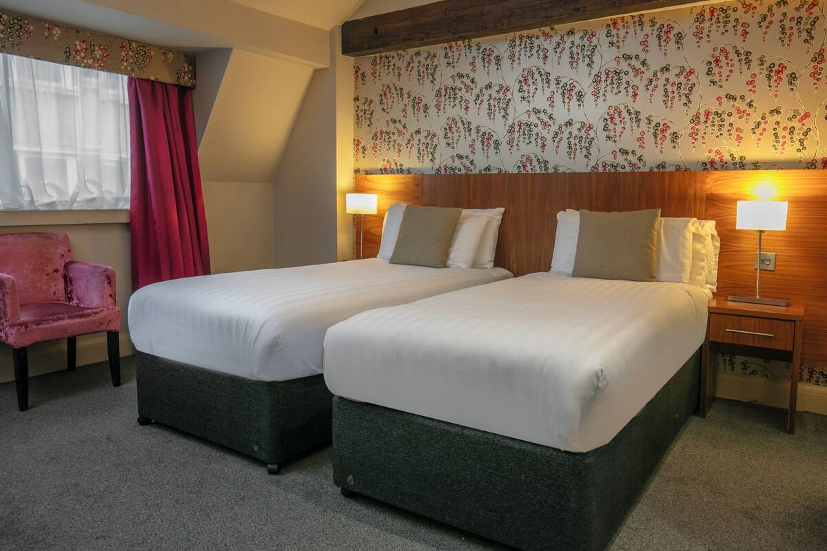 A hotel room with two beds, a wooden headboard, a floral wallpaper, a pink chair, and a window with red curtains.
