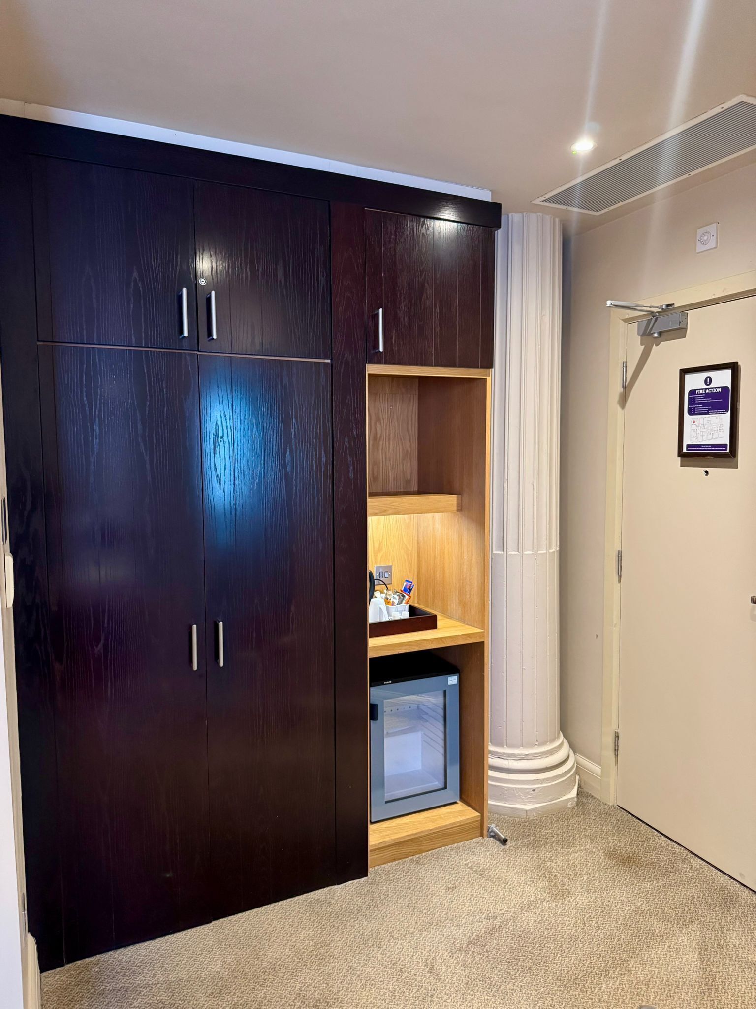 Una habitación con un armario de madera oscura, un pequeño estante con objetos, un cuadro en la pared y una puerta con señal de evacuación.