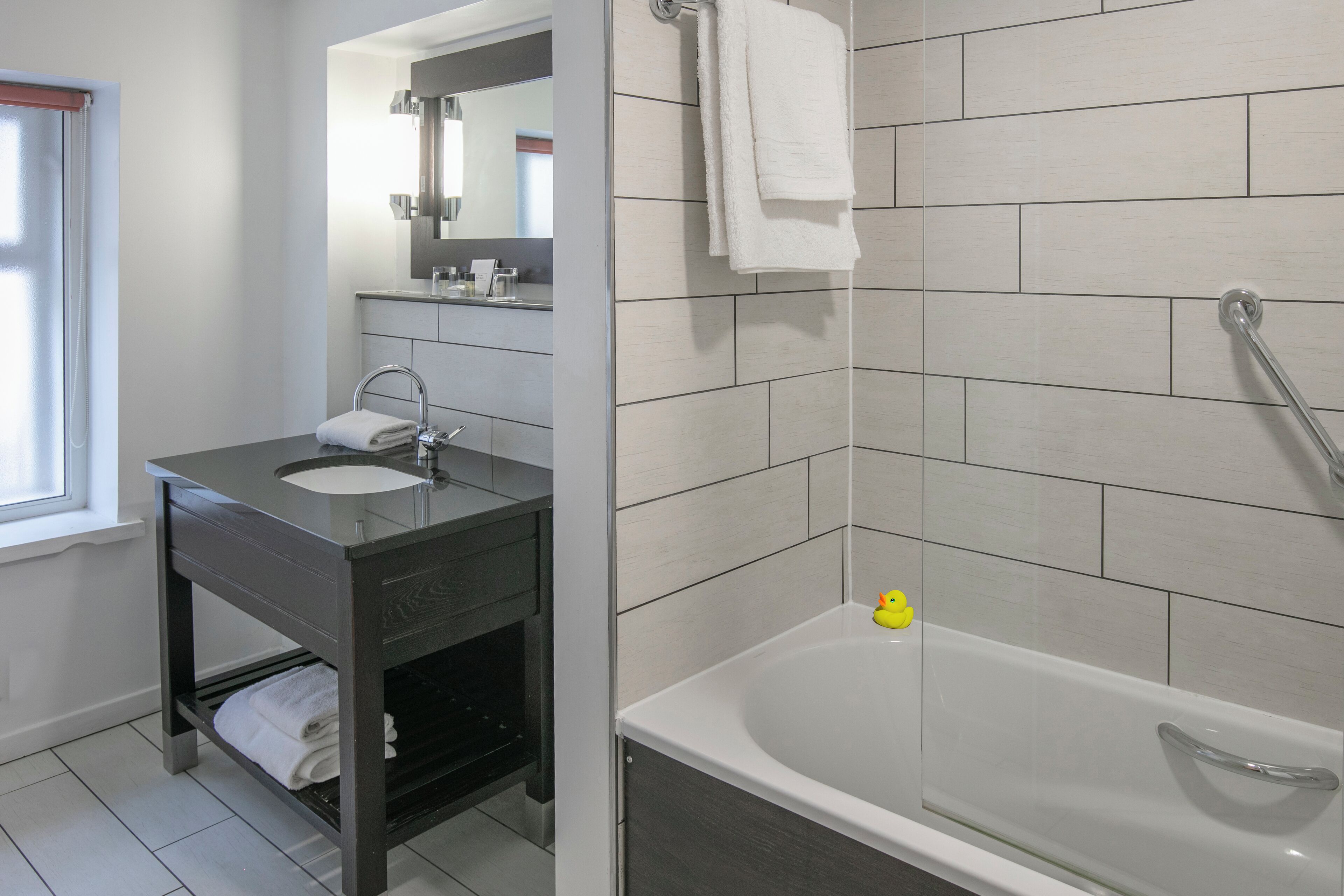 A modern bathroom with a dark vanity, a white sink, a mirror, and a bathtub.