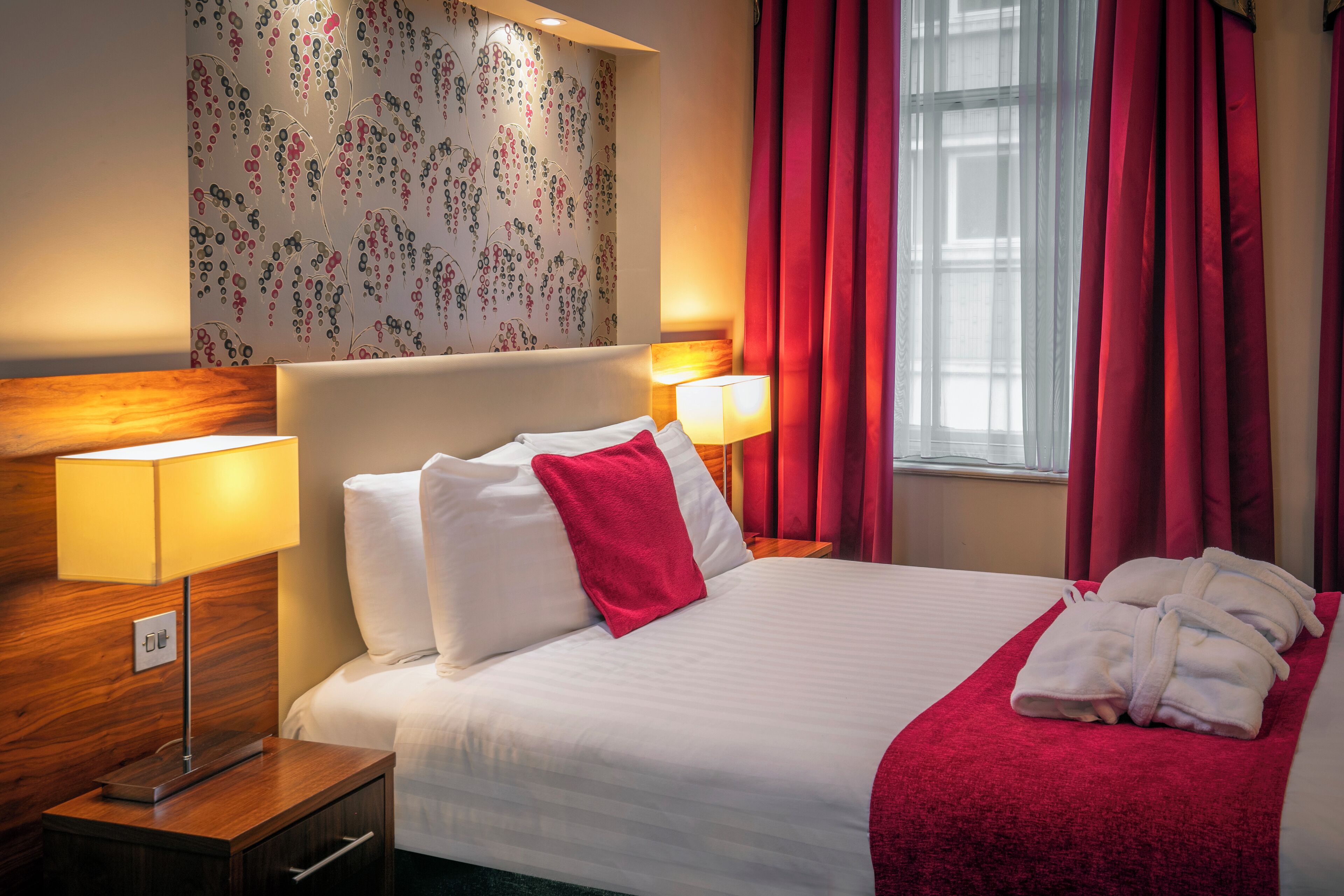 A hotel room with a neatly made bed, two bedside lamps, a wooden nightstand, and a window with red curtains.
