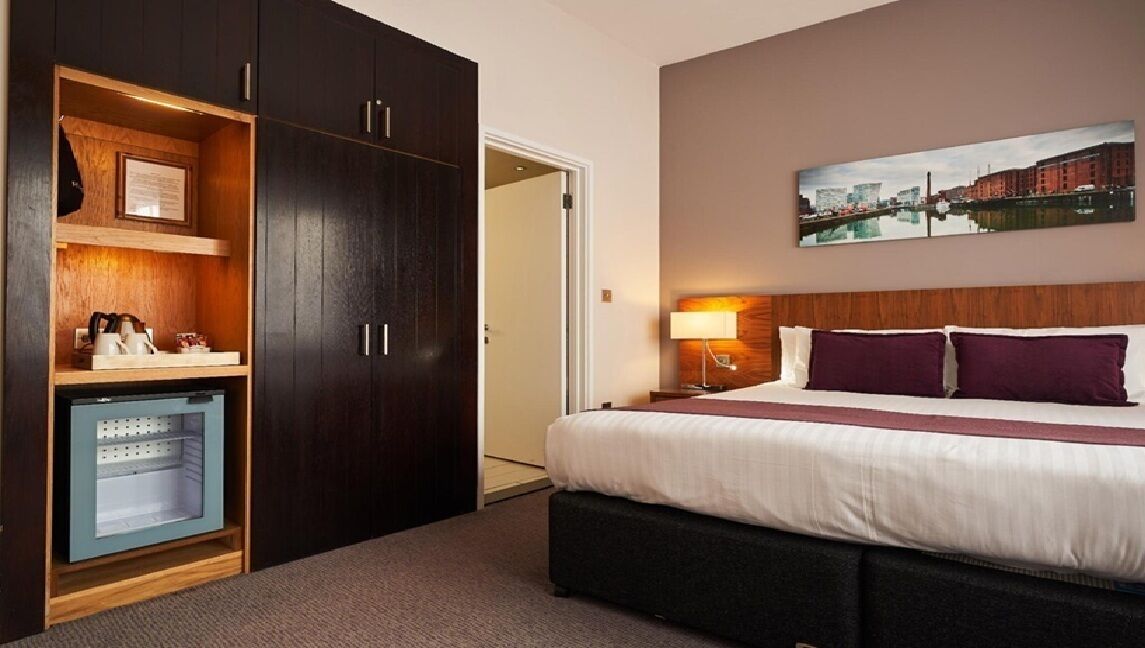 Ein Hotelzimmer mit einem großen Bett, einem Schrank mit Spiegel, einem kleinen Schreibtisch mit Lampe und einem Stadtbild an der Wand.