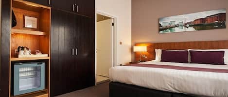 Ein Hotelzimmer mit einem großen Bett, einem Schrank mit Spiegel, einem kleinen Schreibtisch mit Lampe und einem Stadtbild an der Wand.