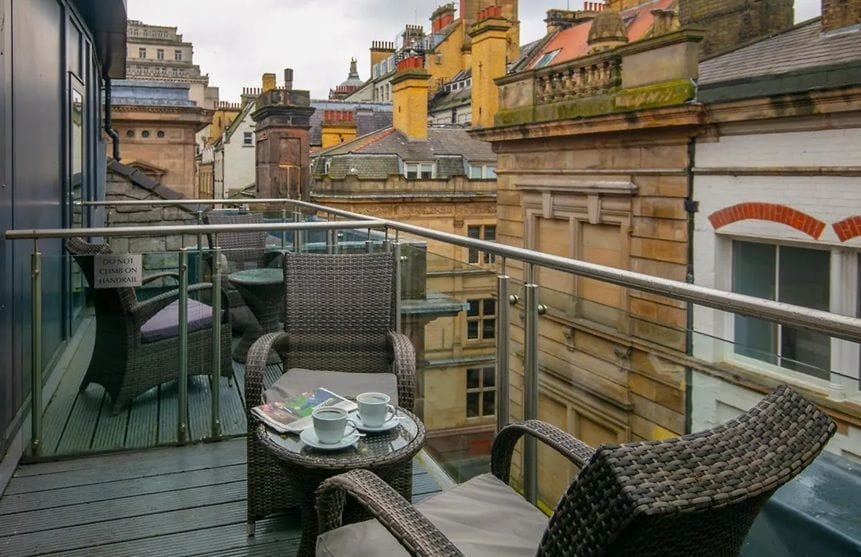 A balcony with wicker furniture and a table, overlooking a cityscape with historic buildings.