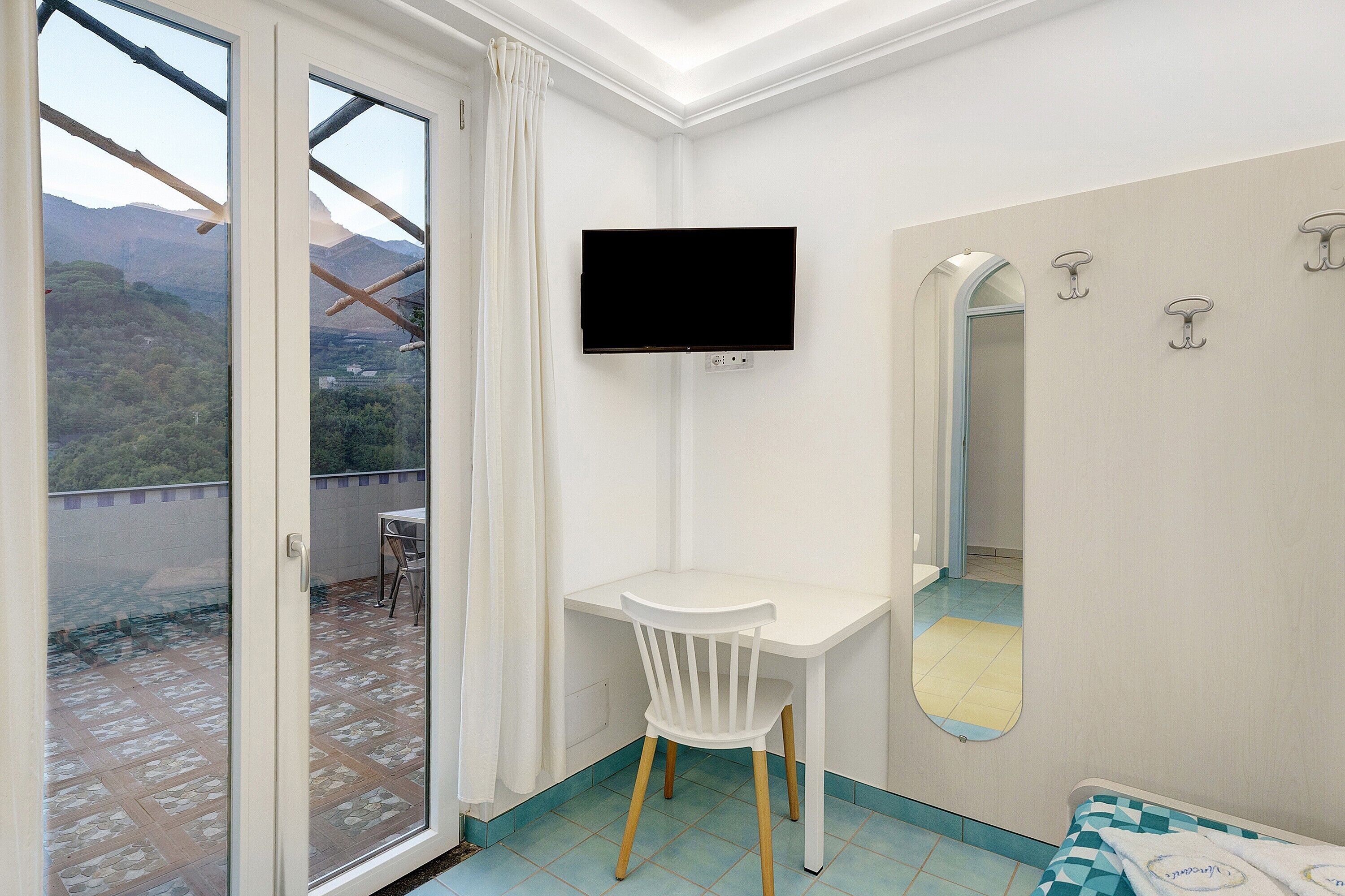 A room with a white desk, a TV mounted on the wall, a mirror, and a view of the outdoors.