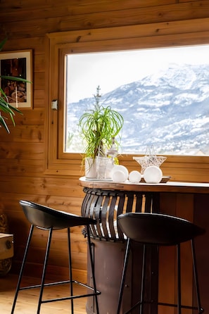 A wooden cabin interior with a wooden table, two black chairs, a potted plant, and a window with a snowy mountain view. - Guesthouse 'Chambre Granon Au Datcha' with Mountain View, Private Terrace and Wi-Fi (Briançon)