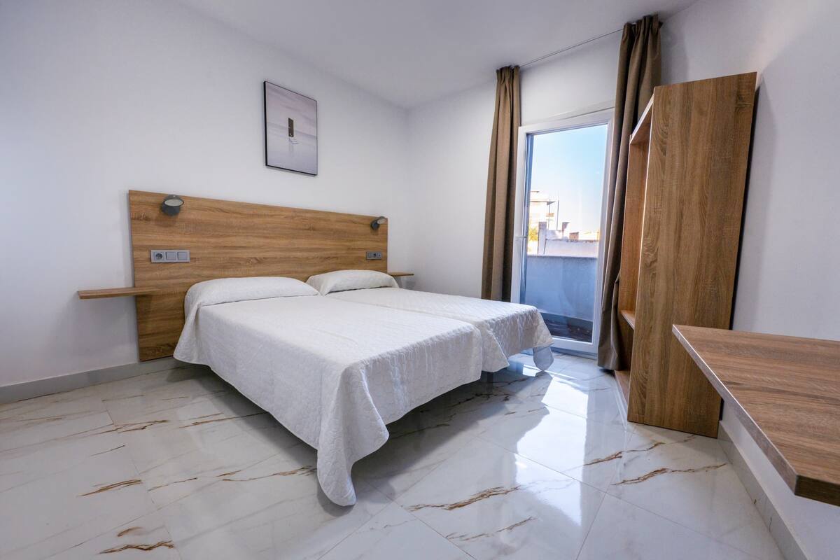Une chambre d’hôtel avec une tête de lit en bois, un lit avec une literie blanche, une petite table de chevet et une vue sur la ville par une grande fenêtre.