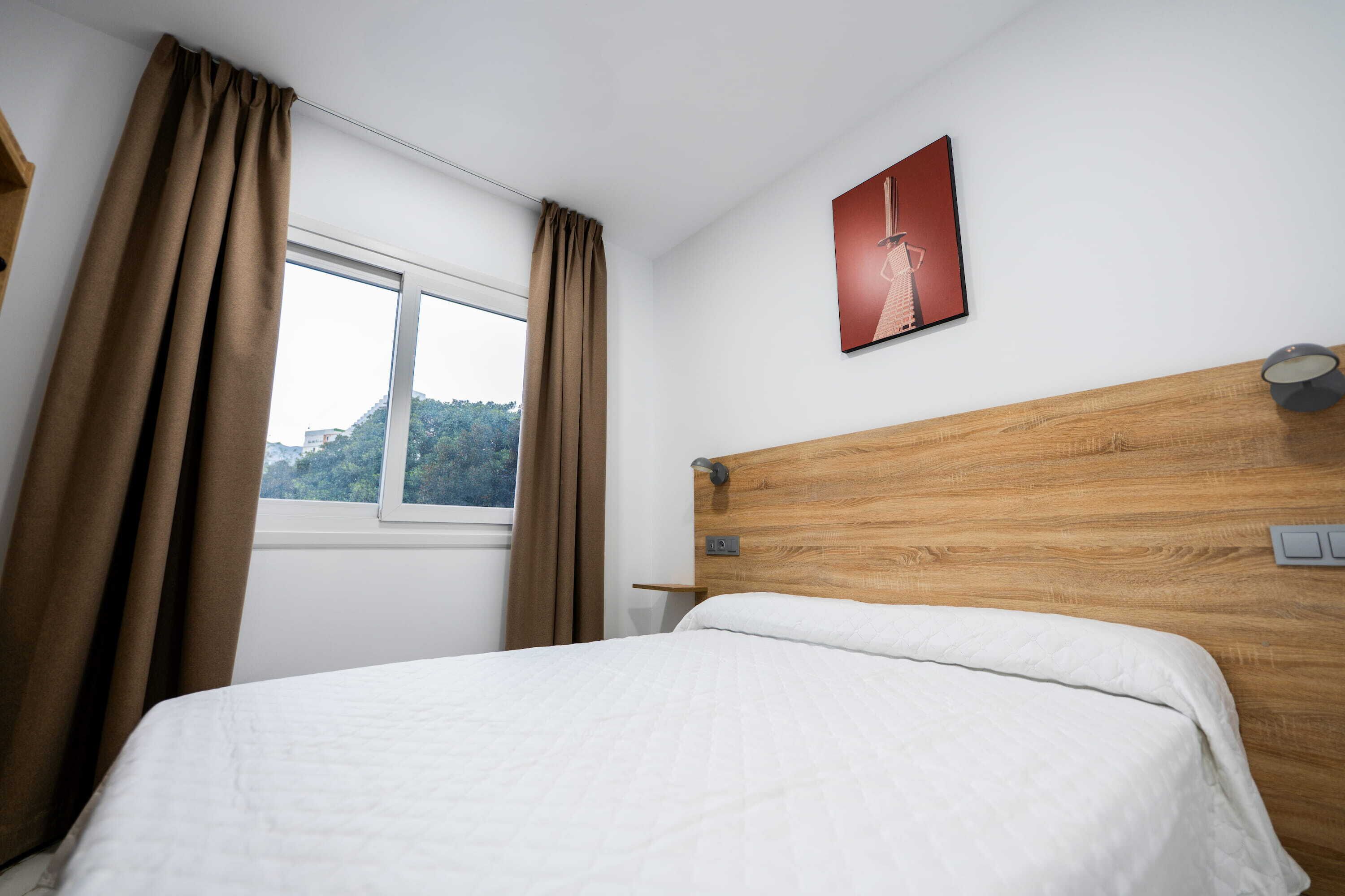 A hotel room with a bed, a window with curtains, a wooden headboard, a wall-mounted light, and a framed picture.