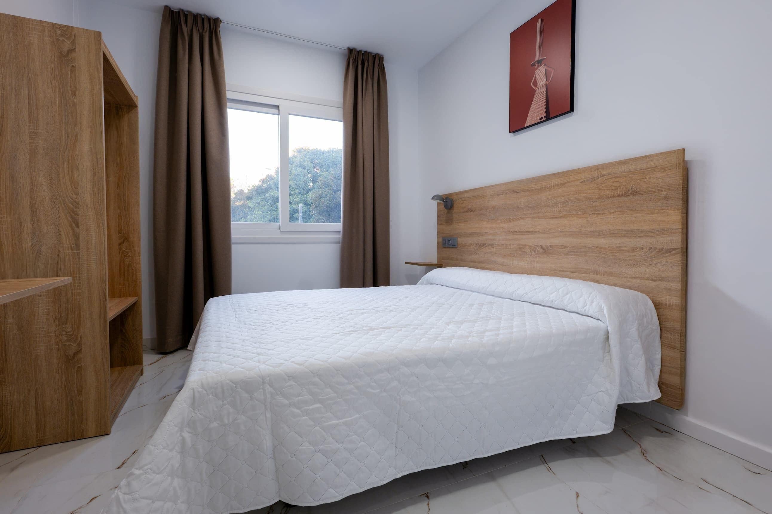 A bedroom with a wooden headboard, a white quilted bedspread, a wardrobe, and a window with curtains.