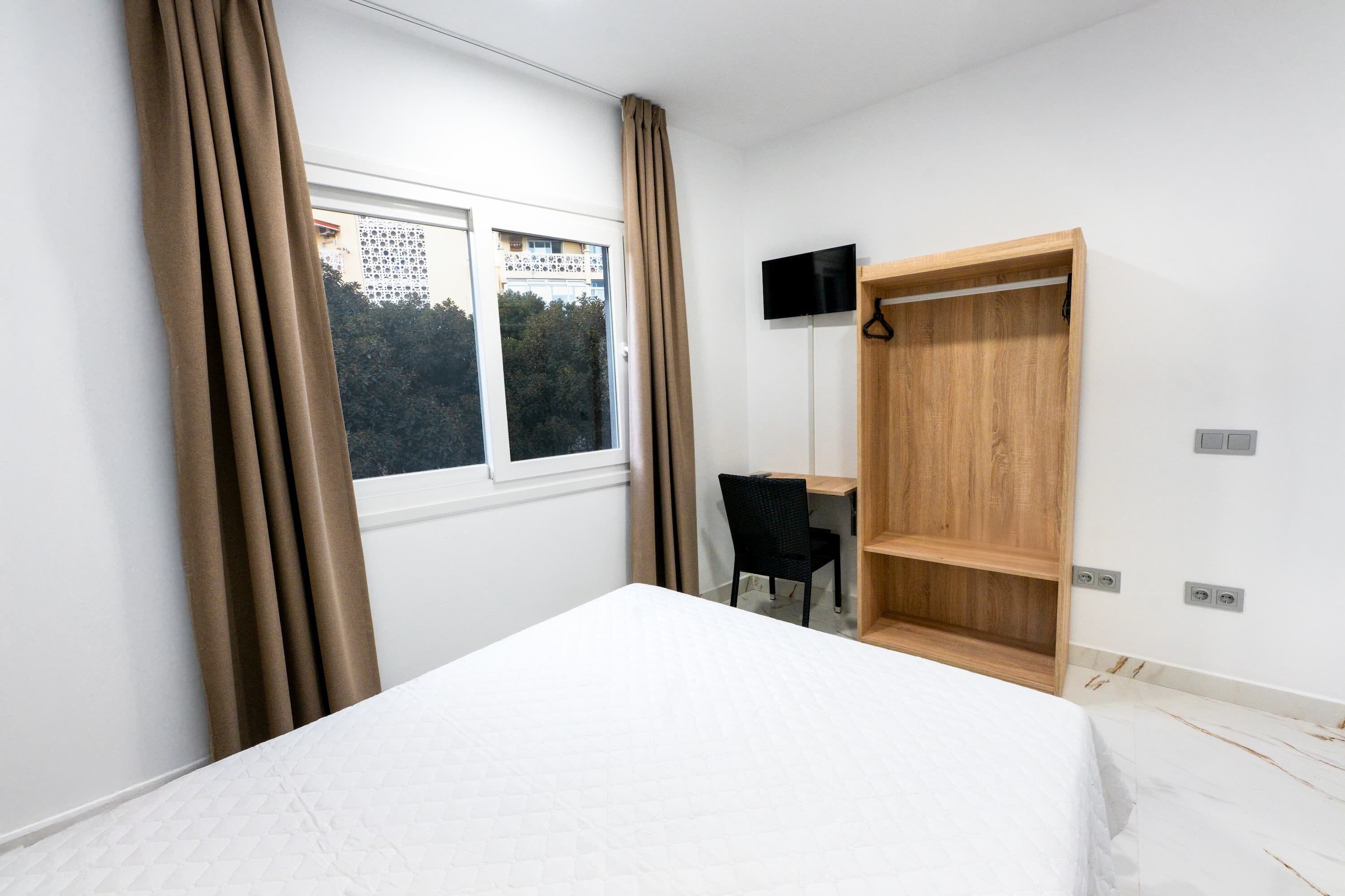 A bedroom with a bed, a desk, a chair, a wardrobe, and a window with curtains.