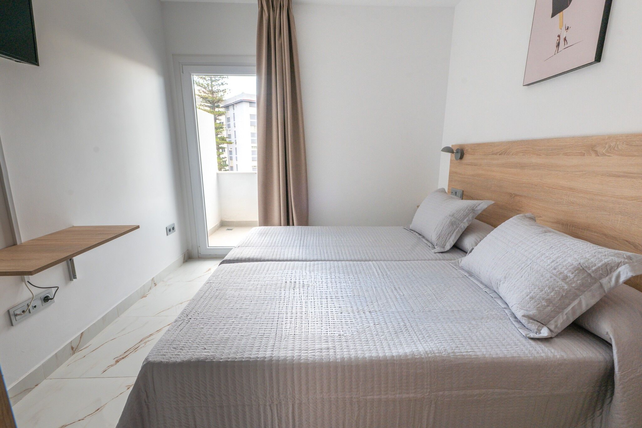 Une chambre avec deux lits, une tête de lit en bois, une table de chevet, une fenêtre avec des rideaux et une vue sur les bâtiments à l’extérieur.