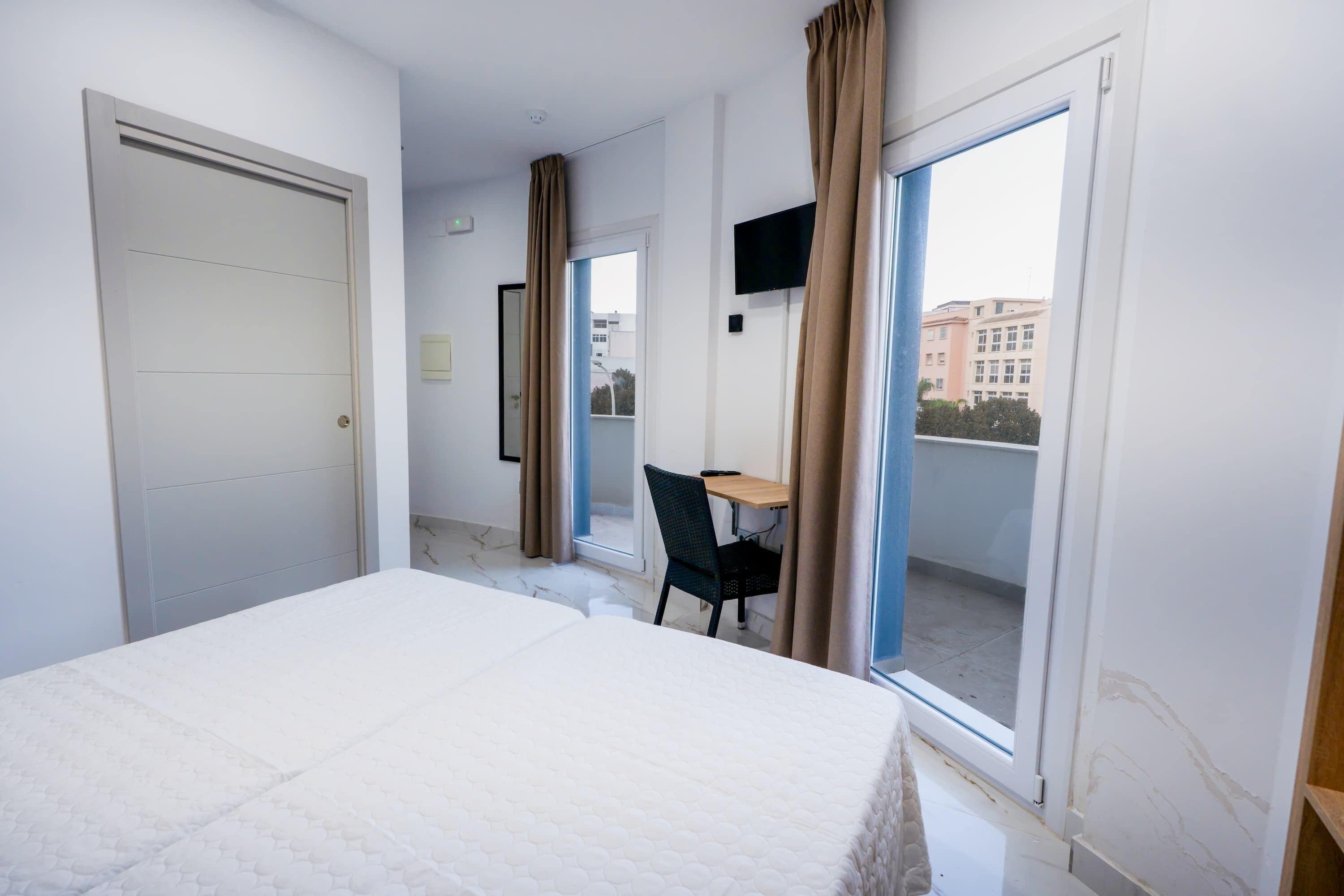 Una habitación de hotel con cama, escritorio, silla, televisor y balcón con cortinas.