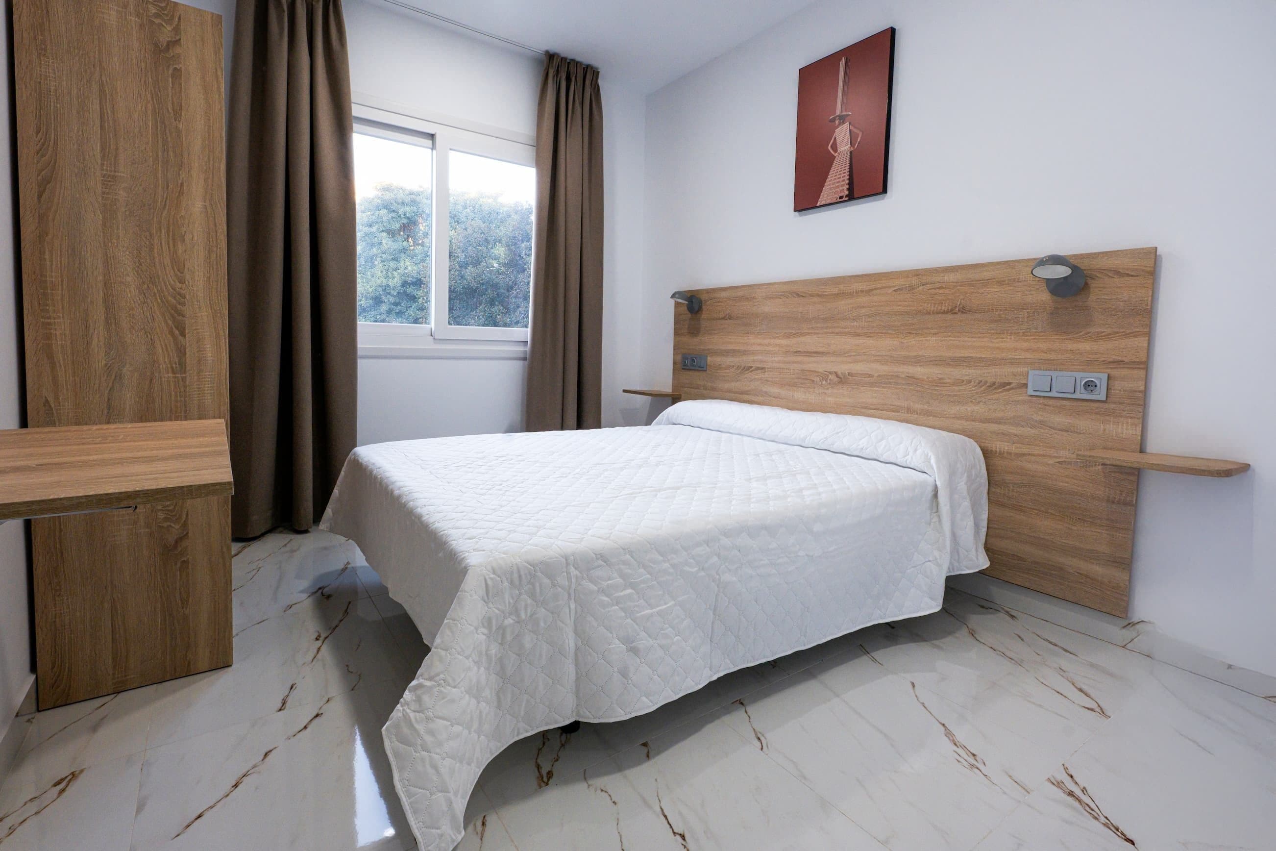 A bedroom with a wooden headboard, a white bedspread, a window with curtains, and a wall-mounted light fixture.