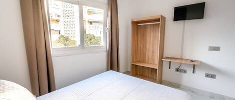 A bedroom with a bed, a window with curtains, a wooden wardrobe, a TV mounted on the wall, and a shelf.