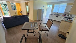 Un espacio de estar con sofá, una mesa y dos sillas, una cocina con armarios y un refrigerador, y vistas al verdor exterior.