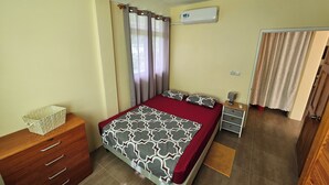 Un dormitorio con cama, cómoda de madera, mesita de noche, aire acondicionado y puerta que da a otra habitación.