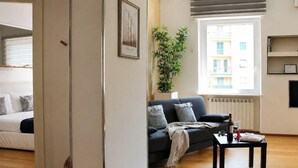 Un salon avec un canapé, une table basse sur laquelle se trouve une bouteille, une plante en pot et une fenêtre donnant sur des immebles.