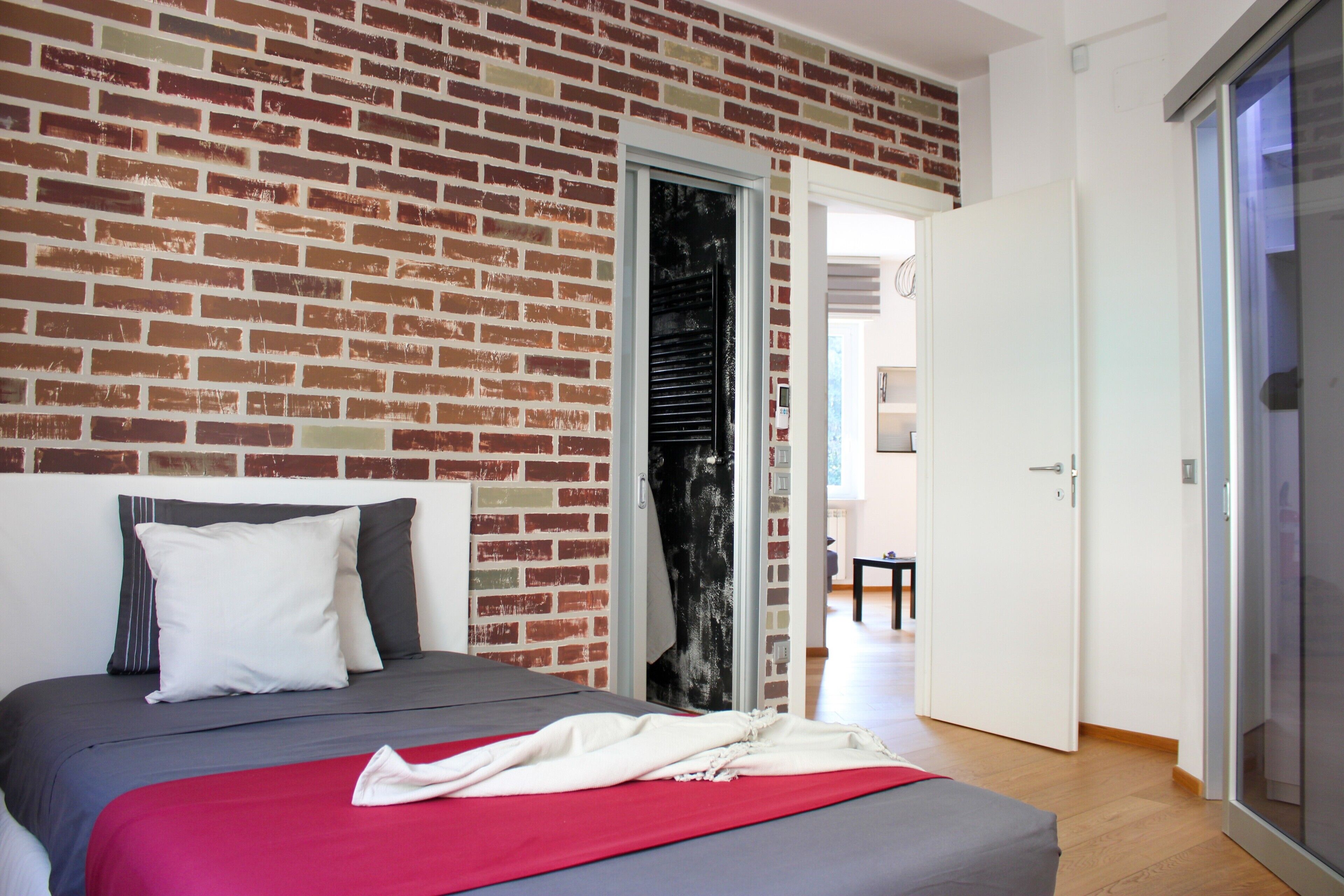 Ein Schlafzimmer mit Backsteinwand, einem Bett mit grauer Bettwäsche, einem weißen Kopfkissen und einer roten Decke.