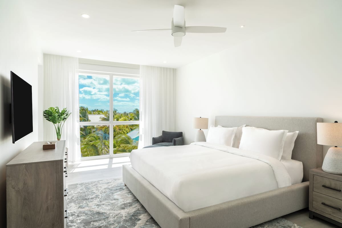 A modern hotel room with a large bed, a ceiling fan, a TV, a nightstand, and a view of a pool and palm trees.